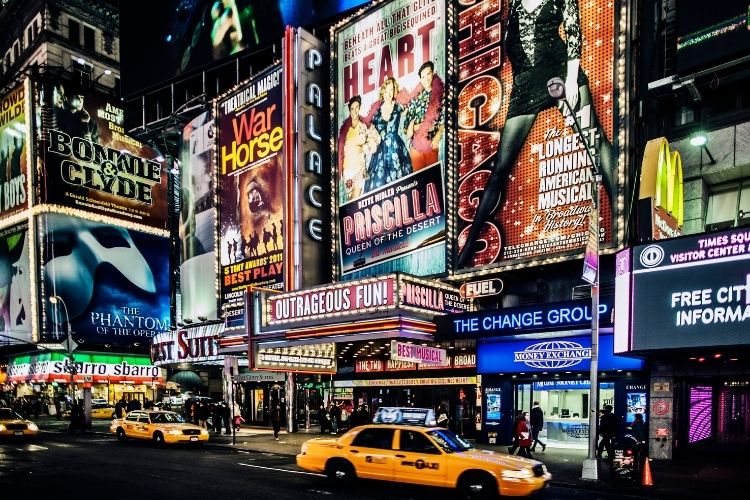 A bustling scene in the heart of New York City, Times Square at night, illuminated by the dazzling, neon-lit billboards and towering skyscrapers. In the foreground, throngs of people navigate the busy intersection, their silhouettes cast against the vibrant backdrop. Capture the iconic architecture, from the grand, Art Deco buildings to the sleek, modern high-rises, all bathed in a warm, golden glow. Convey the energy and excitement of this iconic urban destination, where the bright lights and lively atmosphere create a captivating and visually stunning experience.